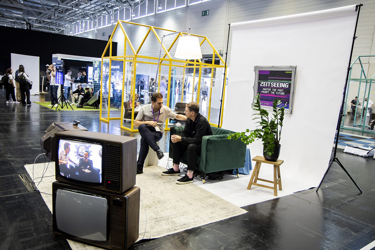 The Zeitseeing (“time seeing”) installation by Jung von Matt at DMEXCO 2024 Two men sitting next to each other in a hall with a cathode ray tube television in front of them.
